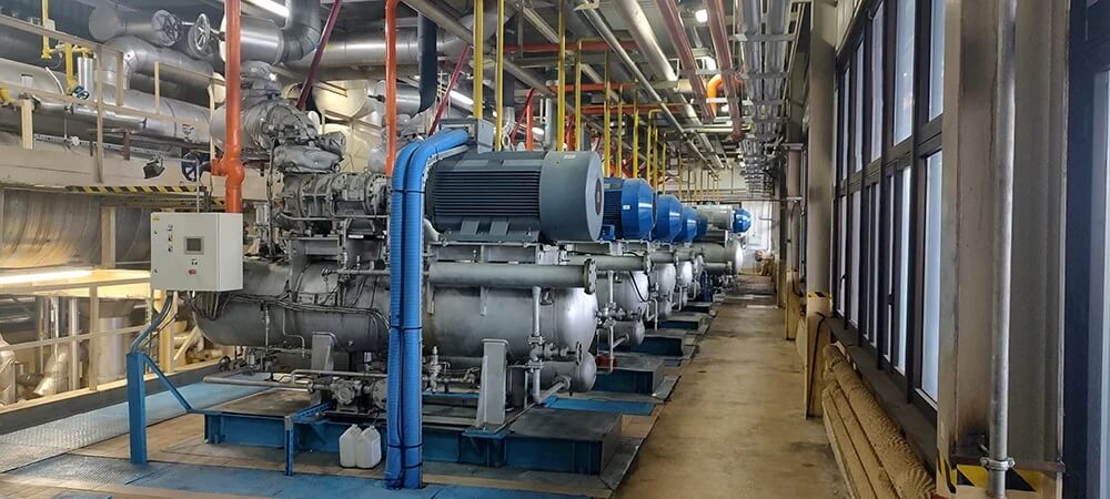 Zdjęcie przedstawia przemysłowe wnętrze z szeregiem dużych sprężarek lub pomp ustawionych na niebieskich platformach. Każde urządzenie wyposażone jest w silnik elektryczny z widocznymi żebrami chłodzącymi, połączony sprzęgłem z systemem rur i zaworów. Nad instalacją rozciąga się gęsta sieć rur, przewodów i kanałów, świadcząca o złożoności systemu transportu cieczy lub gazu. Po lewej stronie znajduje się biały panel sterowania, a po prawej duże okna wpuszczają naturalne światło, rozjaśniając przestrzeń. 