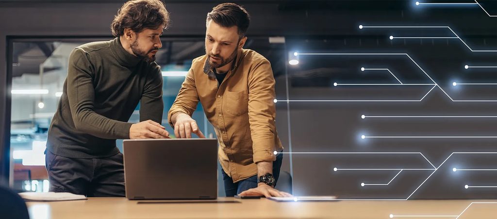 Na zdjęciu widać dwóch mężczyzn analizujących dane przy laptopie. Pracują przy stole i wspólnie omawiają informacje wyświetlane na komputerze. W tle znajduje się nowoczesne, biurowe wnętrze, a całość uzupełniają graficzne linie przypominające schematy obwodów elektronicznych, nawiązujące do technologii, automatyki i cyfryzacji.