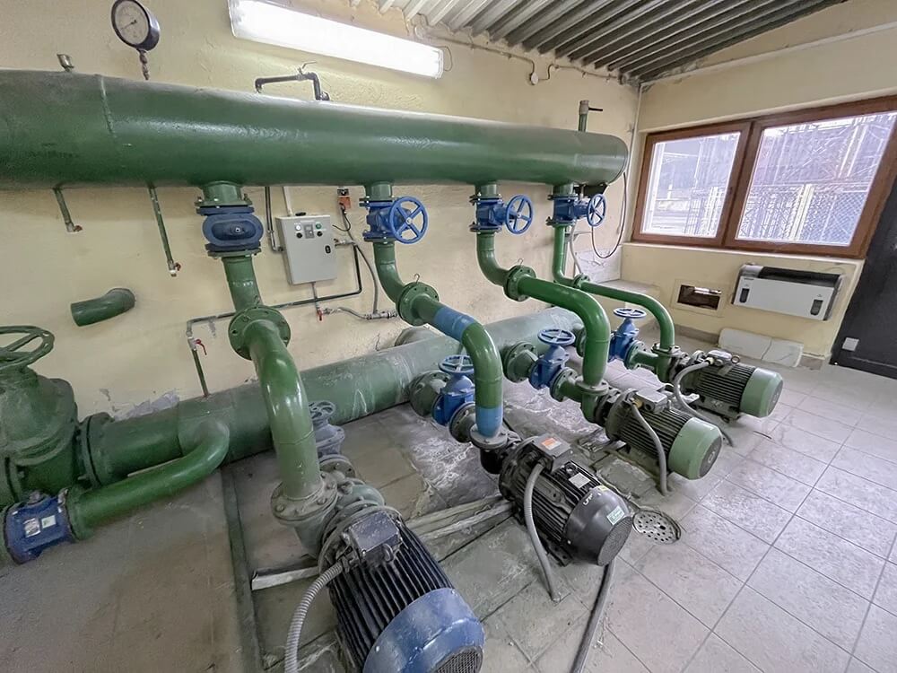 Na zdjęciu widoczna jest techniczna instalacja rurowa z układem pomp i armatury przemysłowej. W pomieszczeniu znajduje się duży, zielony zbiornik lub kolektor połączony z siecią rurociągów, zaworów odcinających i pomp elektrycznych. Na instalacji widoczny jest również manometr wskazujący ciśnienie oraz lokalny panel sterowania.
